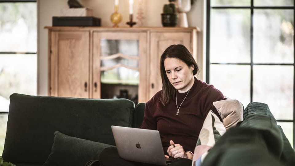 En kvinna sitter i soffan och jobbar med sin dator i knät. 