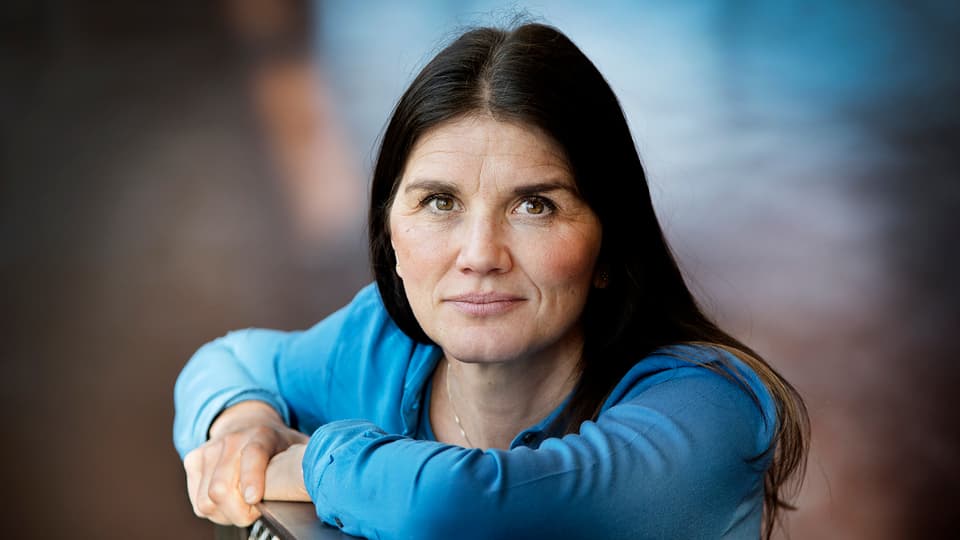 A woman with dark long hair, brown eyes and a blue sweater is leaning against the back of a chair and looking into the camera.